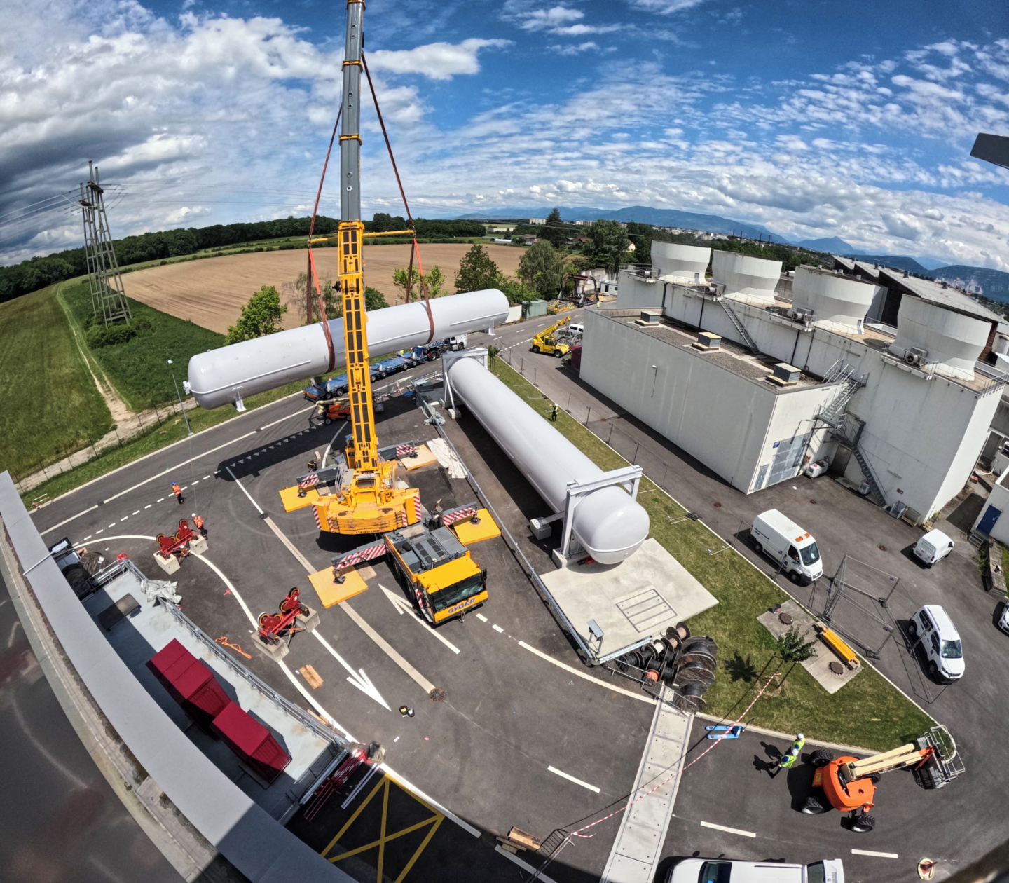 View of two helium tanks (huge white cylindric metallic tanks). One is installed and the other is lifted over the first by a crane.