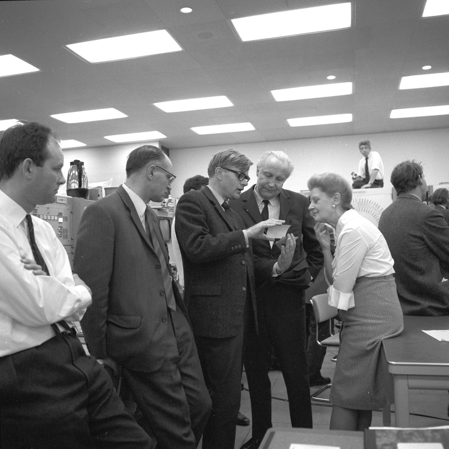 Man showing man and woman a photo in a control room full of people