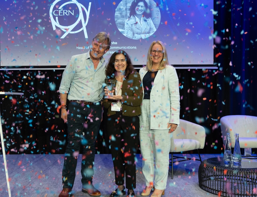 1 man and 2 women, side to side, smiling at the camera. The woman in the middle has an award in the hands and there's confetti tumbling in front of them. 